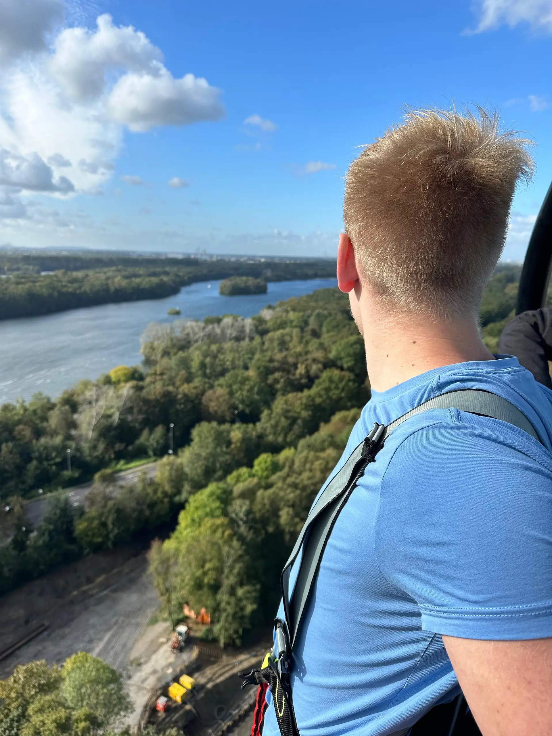 Bungeespringer mit kurzem Haar schaut aus großer Höhe auf einen ausgedehnten Wald und ein Gewässer.